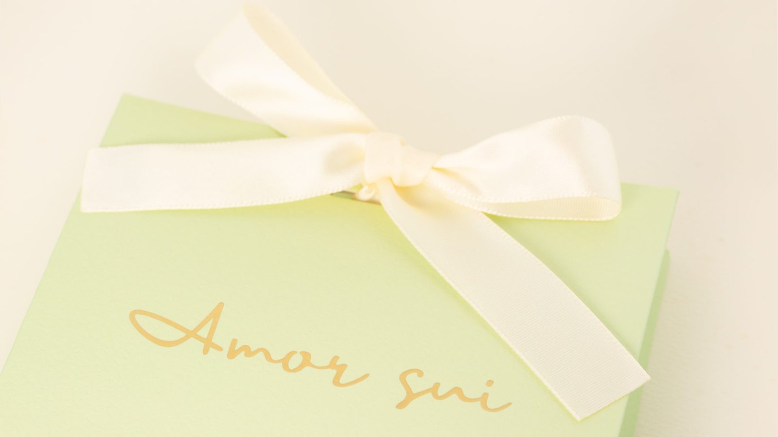 Green gift box with "Amor Sui" written in elegant gold script and adorned with a white satin ribbon bow.